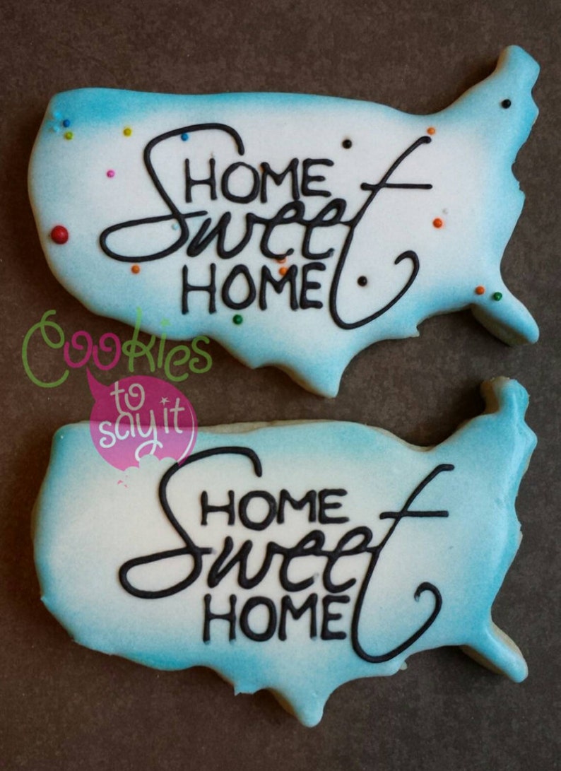 May include: Two blue and white sugar cookies decorated with a white background and black icing. The cookies are shaped like the United States of America and have the words "Home Sweet Home" written on them.