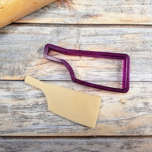 Peut inclure: Un emporte-pièce en plastique violet en forme de bouteille de vin sur une surface en bois avec un morceau de pâte découpé à l'aide de l'emporte-pièce.