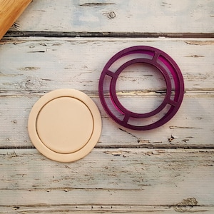 May include: A close-up of a purple plastic cookie cutter shaped like a ring with a smaller ring inside. A beige circle of dough is next to the cookie cutter.