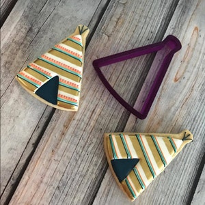 May include: Two decorated sugar cookies in the shape of a teepee, with a purple plastic cookie cutter in the shape of a teepee. The cookies are decorated with gold, white, and orange stripes, and a blue triangle in the center.