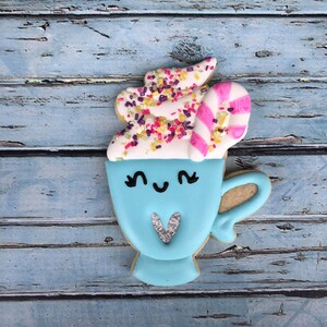 Puede incluir: Una galleta de az&uacute;car azul y blanca decorada para parecer una taza de chocolate caliente con crema batida, chispas y un bast&oacute;n de caramelo. La taza tiene una carita linda con ojos y un coraz&oacute;n.
