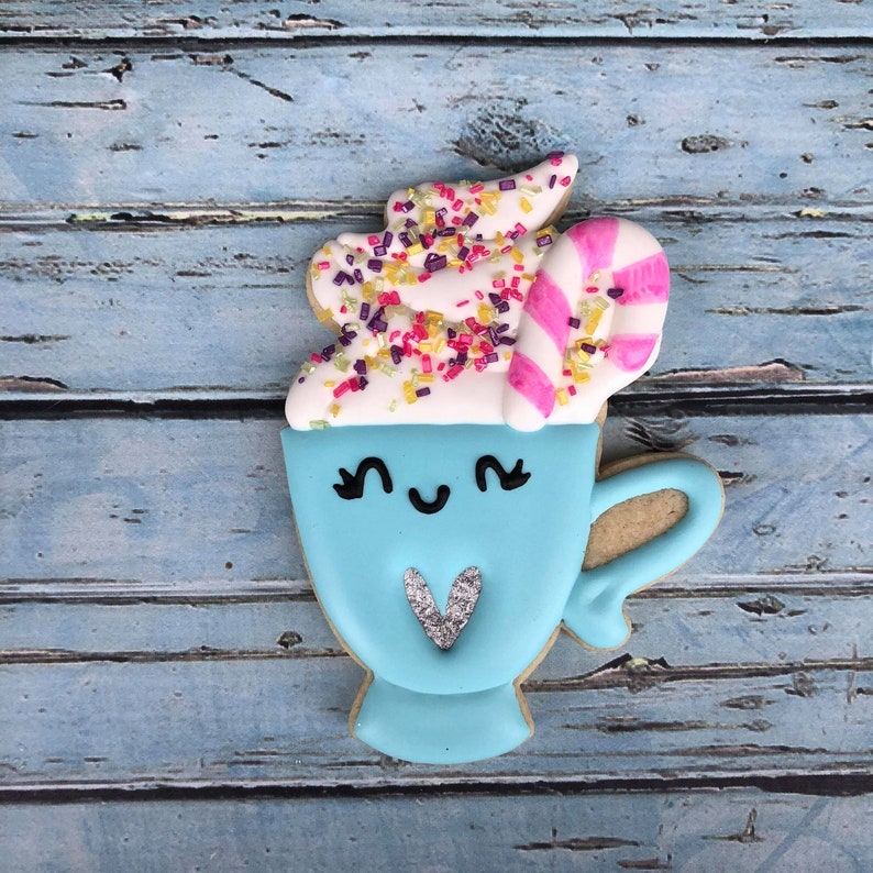 Puede incluir: Una galleta de az&uacute;car azul y blanca decorada para parecer una taza de chocolate caliente con crema batida, chispas y un bast&oacute;n de caramelo. La taza tiene una carita linda con ojos y un coraz&oacute;n.