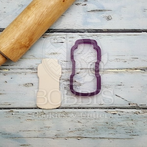 May include: A purple plastic cookie cutter shaped like a water bottle, next to a wooden rolling pin and a white cookie cutout of the same shape.