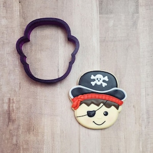 May include: Purple plastic cookie cutter shaped like a pirate's head with an eye patch and a hat with a skull and crossbones. A decorated sugar cookie with the same design is next to the cutter.