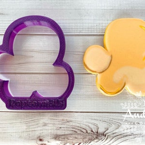 May include: Purple plastic cookie cutter shaped like a popcorn kernel with the words "Pop It Up" on the bottom. Next to the cutter is a yellow popcorn kernel cookie.