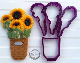 May include: Purple plastic cookie cutters shaped like a vase and three sunflowers. The cutters are on a white surface.