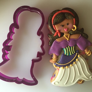 May include: Purple and white cookie cutter in the shape of a woman wearing a purple and gold dress. The cookie cutter is next to a decorated cookie of a woman wearing a purple and gold dress.