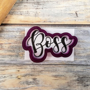 May include: Purple plastic cookie cutter in the shape of the word "Boss" in a cursive font. The cutter is on a white piece of paper and a wooden rolling pin is in the background.