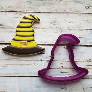 May include: A purple plastic witch hat cookie cutter and a decorated witch hat cookie with yellow and black stripes and a black buckle.