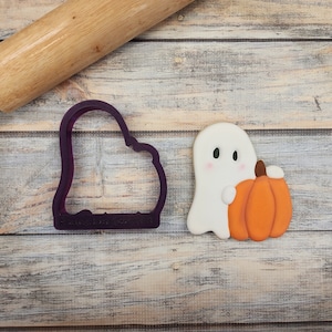 May include: A white ghost cookie with a small orange pumpkin in its hand. A purple plastic cookie cutter in the shape of a ghost is also shown.