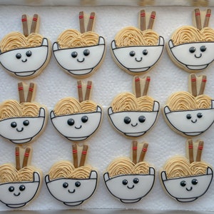 May include: Twelve decorated sugar cookies shaped like bowls of ramen noodles with chopsticks. Each cookie has a smiling face and is decorated with brown, white, and red icing.