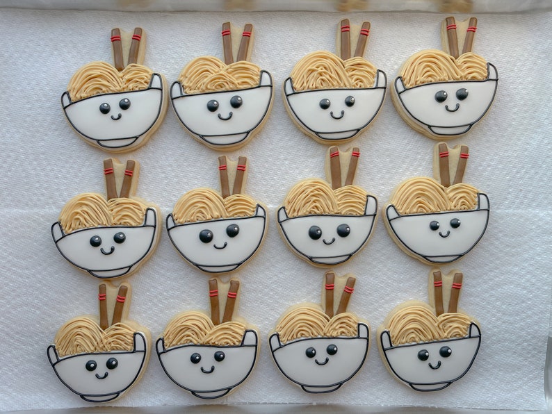 May include: Twelve decorated sugar cookies shaped like bowls of ramen noodles with chopsticks. Each cookie has a smiling face and is decorated with brown, white, and red icing.