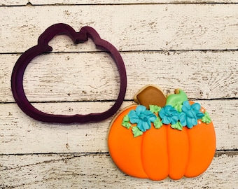May include: A purple pumpkin cookie cutter and a decorated pumpkin cookie with teal and green icing.