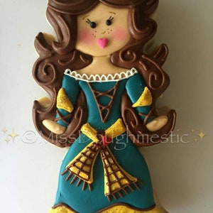 May include: A decorated sugar cookie shaped like a woman with brown hair, a blue dress, and a yellow sash. The cookie has a pink heart on its face and is decorated with brown icing.