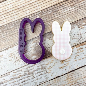 May include: Purple plastic bunny cookie cutter with the text "Bunny 10 4" on the side, next to a pink and white gingham bunny cookie.