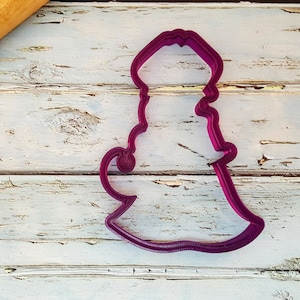 May include: A purple plastic cookie cutter shaped like a genie in a bottle. The cookie cutter is on a white wooden surface.