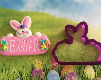 May include: Pink cookie decorated with a white bunny peeking over the top and the words "Happy Easter" in white icing. The cookie is decorated with colorful sprinkles. A purple plastic cookie cutter in the shape of a bunny is also shown.