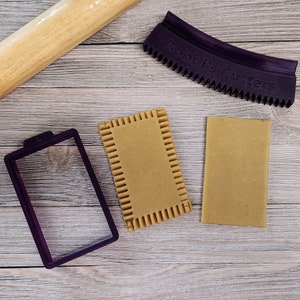 May include: Two brown cookie dough rectangles, a purple plastic cookie cutter, and a wooden rolling pin on a light wood surface. The cookie cutter has the text "Bobbie's Cutters" on it.