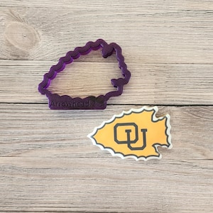 May include: A purple arrowhead-shaped cookie cutter with the text "Arrowhead 2.4" and a yellow and black arrowhead-shaped sticker with the letters "CU" on a wooden surface. The cookie cutter has a wavy edge.