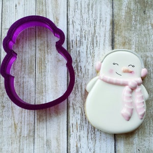 May include: Purple plastic snowman cookie cutter with a white and pink iced snowman cookie. The snowman has a pink scarf and earmuffs.