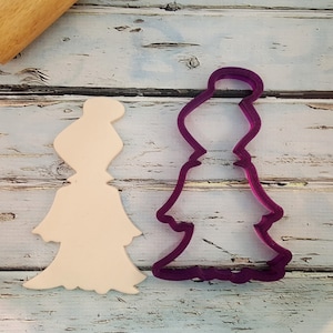 May include: A white plastic cookie cutter in the shape of a woman wearing a dress. The cookie cutter is on a white wooden surface. A purple plastic cookie cutter in the same shape is next to it.