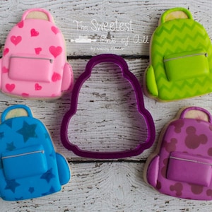 May include: Five decorated sugar cookies shaped like backpacks. The cookies are decorated with different patterns, including hearts, stars, zigzags, and Mickey Mouse heads. The cookies are arranged on a white wooden surface. The text "The Sweetest One of All" is visible in the background.