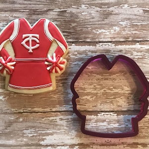 May include: A red and white cheerleader uniform cookie cutter. The cutter is made of purple plastic and is shaped like a cheerleader outfit with pom-poms.
