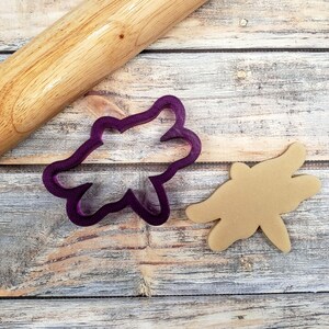 May include: Purple plastic cookie cutter shaped like a flower, next to a brown cookie cut out in the same shape on a wooden surface. A wooden rolling pin is also in the image.