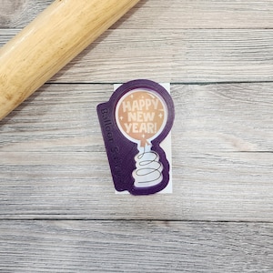 May include: Purple plastic cookie cutter shaped like a balloon with the text "Happy New Year!" inside. The cookie cutter is labeled "Balloon Stamper".