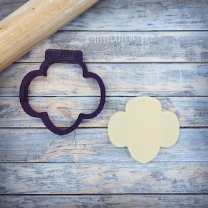 May include: A purple plastic trefoil-shaped cookie cutter with the text "Trefoil 4". A cut-out cookie in the same shape is next to the cutter. A wooden rolling pin is in the background. The items are on a wooden surface.