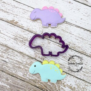 Peut inclure: Trois biscuits en forme de dinosaure, un violet, un bleu et un emporte-pièce. Le biscuit violet a des points roses et le biscuit bleu a des points jaunes. L'emporte-pièce est violet.
