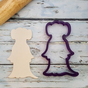 May include: A purple plastic cookie cutter in the shape of a female character from Star Wars. The cutter is on a wooden surface next to a wooden rolling pin.