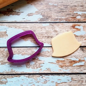 May include: A purple plastic cookie cutter in the shape of a shield, next to a piece of beige fondant cut in the same shape. The items are on a distressed, light blue and brown wooden surface. A wooden rolling pin is in the background.