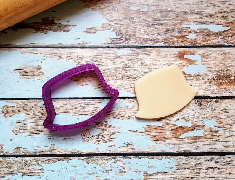 May include: A purple plastic cookie cutter in the shape of a shield, next to a piece of beige fondant cut in the same shape. The items are on a distressed, light blue and brown wooden surface. A wooden rolling pin is in the background.