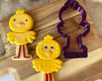 Op de afbeelding: Twee gele kuiken koekjesvormen en een paarse kuiken koekjesvorm op een houten snijplank. De koekjesvormen liggen op een bruine jute ondergrond. Een houten deegroller met de tekst "Drizzle Top Baking" is op de achtergrond.