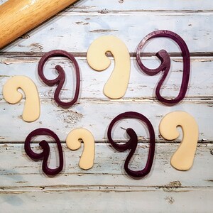 Puede incluir: Un juego de seis cortadores de galletas, tres morados y tres beige, con forma de bast&oacute;n de caramelo. Los cortadores est&aacute;n dispuestos sobre una superficie de madera azul claro.