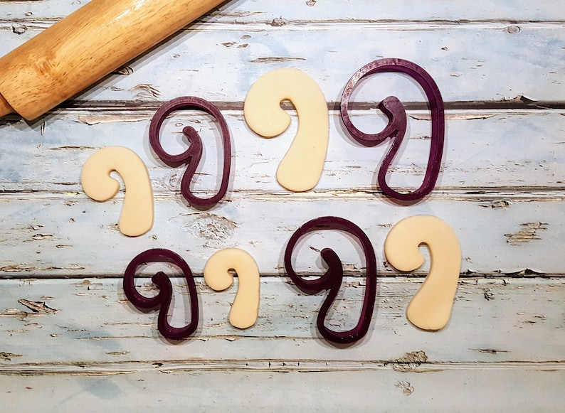 Puede incluir: Un juego de seis cortadores de galletas, tres morados y tres beige, con forma de bast&oacute;n de caramelo. Los cortadores est&aacute;n dispuestos sobre una superficie de madera azul claro.