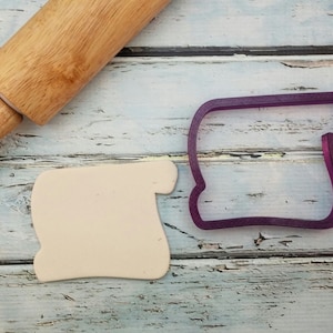 May include: A purple plastic cookie cutter shaped like a slice of bread, next to a piece of white fondant and a wooden rolling pin.