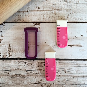 May include: Two pink and white cookie cutters shaped like test tubes. One cutter is purple plastic and the other is a cookie.