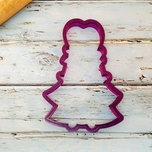 May include: A purple plastic cookie cutter shaped like a girl wearing a dress. The cookie cutter is on a white wooden surface.