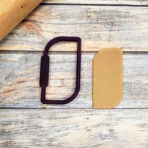 May include: A purple plastic cookie cutter shaped like a rounded rectangle with a handle. A brown piece of fondant is next to the cutter.
