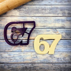 May include: A purple plastic cookie cutter in the shape of the number 67, next to a baked cookie in the same shape. The cookie cutter is on a weathered, light blue and white wooden surface. A wooden rolling pin is in the background.