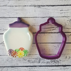 May include: A purple cookie cutter in the shape of a jar with a lid. Next to it is a decorated cookie in the shape of a jar with a lid and colourful candy inside.