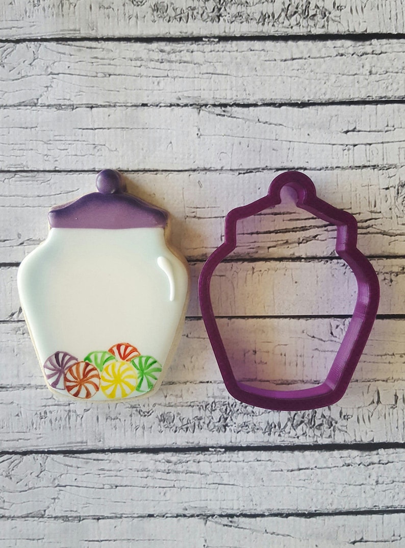 May include: A purple cookie cutter in the shape of a jar with a lid. Next to it is a decorated cookie in the shape of a jar with a lid and colourful candy inside.