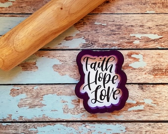 Op de afbeelding: Paarse plastic koekjesvormer met de woorden "Faith Hope Love" in een cursief schrift.