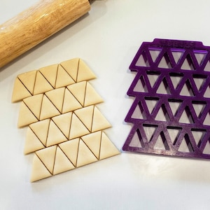 May include: A close-up of a purple triangle cookie cutter and a sheet of dough cut into triangles. The dough is ready to be baked.