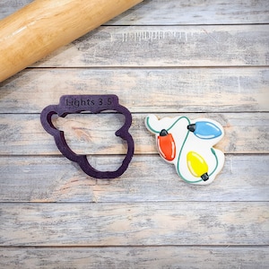 May include: A purple plastic cookie cutter shaped like a string of Christmas lights with the text "Lights 3.5" on it. A decorated sugar cookie with a string of lights design is next to the cutter.