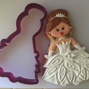 May include: A white cookie decorated with a girl wearing a white dress with a white flower on the bodice. The cookie is next to a purple plastic cookie cutter in the shape of a girl wearing a dress.