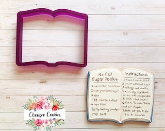 May include: A purple plastic cookie cutter shaped like an open book. The cookie cutter is on a white wooden surface. A handwritten recipe for sugar cookies is visible in the background. The recipe includes ingredients like butter, sugar, eggs, vanilla extract, flour, baking powder, and salt. The recipe also includes instructions for preheating the oven, creaming butter and sugar, adding eggs and vanilla, mixing dry ingredients, chilling the dough, rolling the dough, cutting shapes, and baking the cookies.