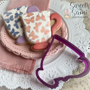May include: Two decorated sugar cookies in the shape of teacups with butterfly designs. A purple plastic cookie cutter in the shape of a teacup is on a white plate with a pink lace border.